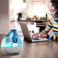 Blue cool mist humidifier on a table beside a woman working on a laptop